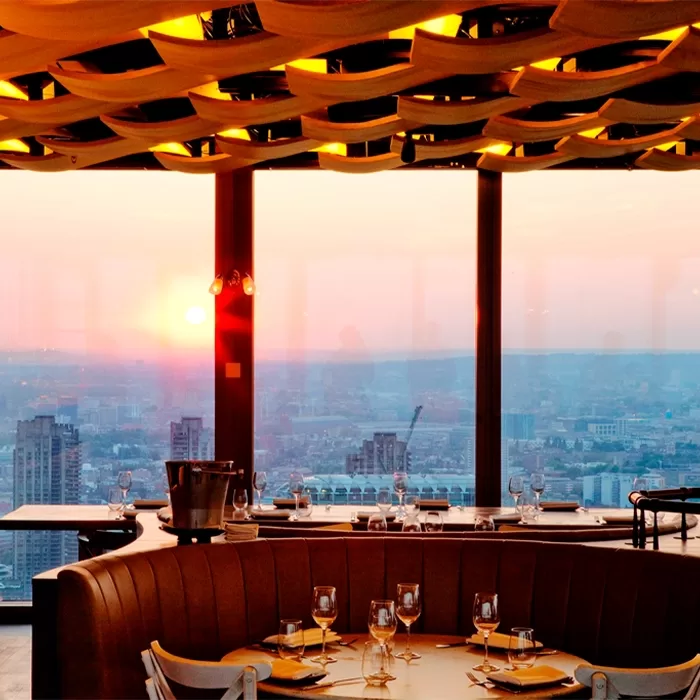 Elegant restaurant with set tables overlooks a city skyline at sunset, seen through large windows. Warm lighting from a decorative ceiling adds to the inviting atmosphere.