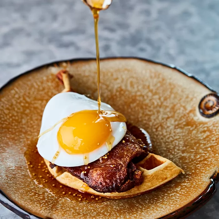 A plate with a waffle topped with crispy fried chicken and a sunny-side-up egg, as syrup is being poured over the dish.