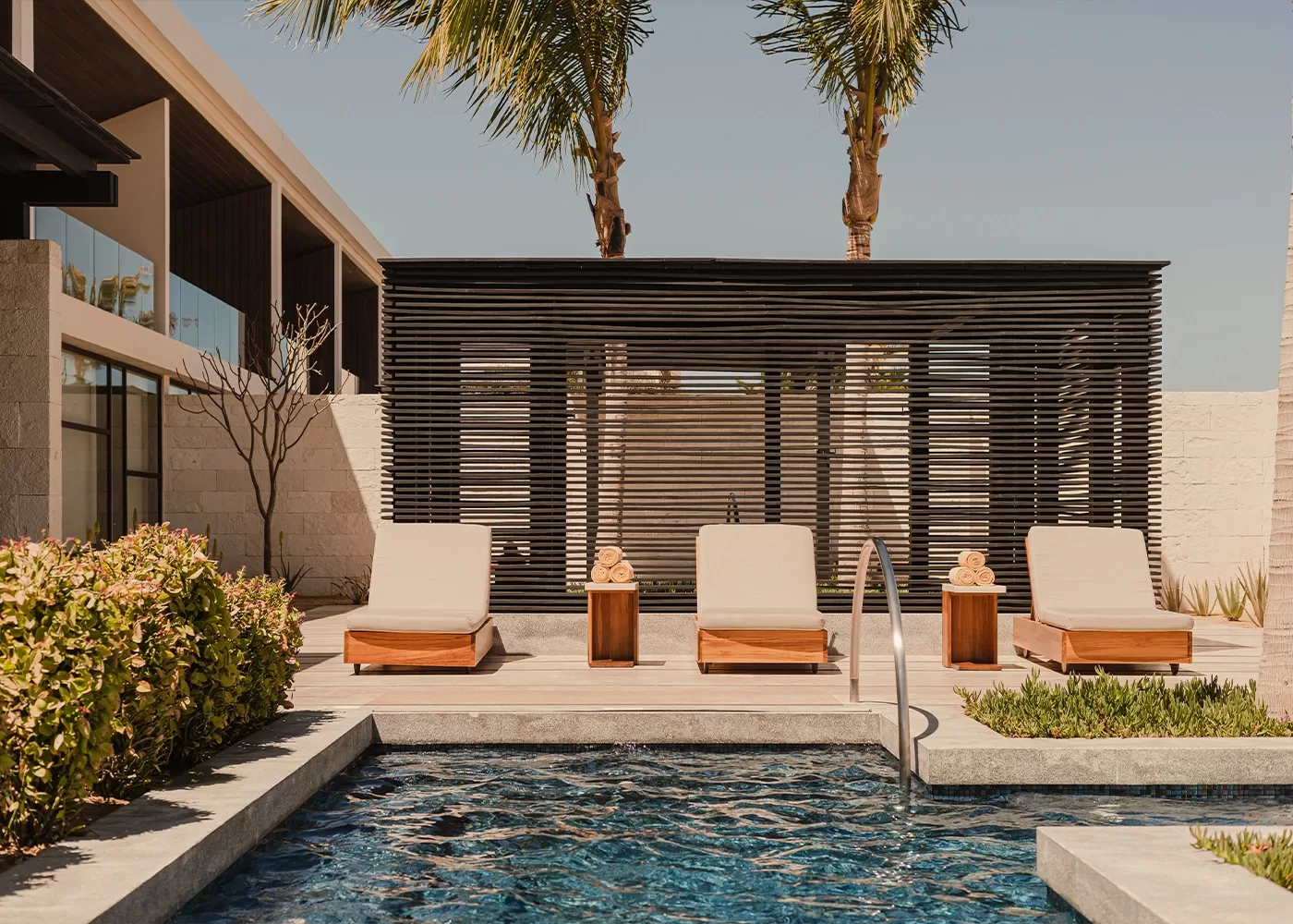 A modern outdoor pool area with three beige lounge chairs and side tables on a wooden deck, shaded by a slatted pergola and two tall palm trees, with clear blue water in the foreground.