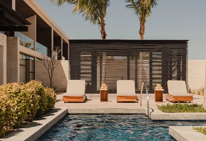 Three lounge chairs with side tables are arranged by a modern pool, with palm trees, greenery, and a minimalist wooden pergola in the background under a clear sky.