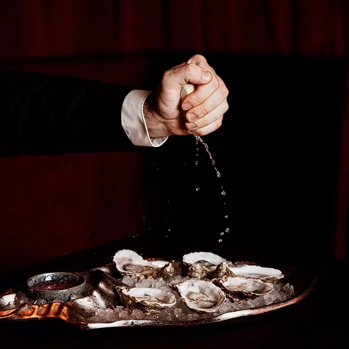 A hand squeezes a lemon over a tray of opened oysters on ice, with droplets of juice falling onto the seafood. The background is dark, drawing focus to the action.