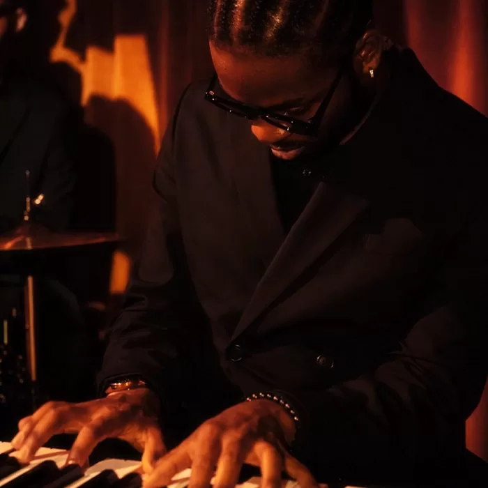 A person wearing dark sunglasses and a black suit plays a keyboard under warm stage lighting, focusing intently on the keys.