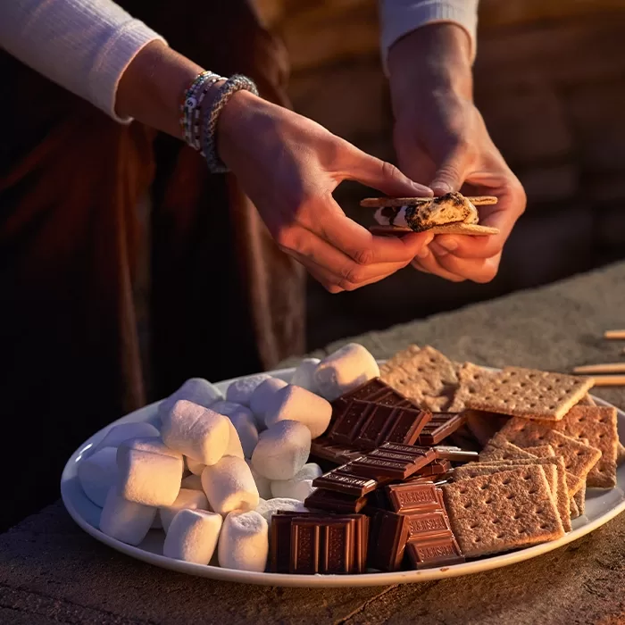 A person assembles a smore above a plate filled with marshmallows, chocolate bars, and graham crackers, set on a stone surface by warm ambient lighting.