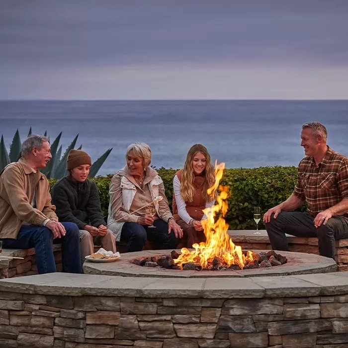 Five people sit around a stone fire pit, smiling and talking, with the ocean and cloudy sky in the background. Everyone is dressed warmly, enjoying an outdoor evening by the fire.