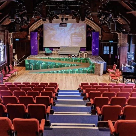 An empty auditorium with red chairs faces a modern stage featuring green geometric designs, chairs, a table, and a large screen displaying presentation content. The venue has wooden floors and arched ceilings.