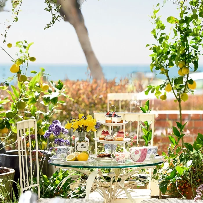 A glass patio table set for tea with floral china, pastries, and a jar of yellow flowers. Surrounded by lush plants, lemons, and two chairs, with the sea and trees visible in the background.