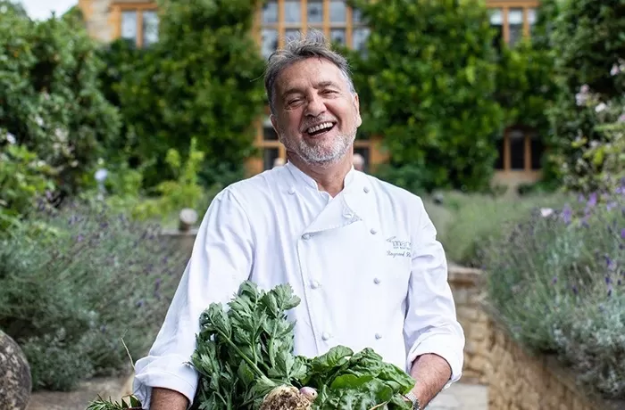 Raymond Blank in his chefs uniform outside of a house carrying a box of vegeatables in a garden