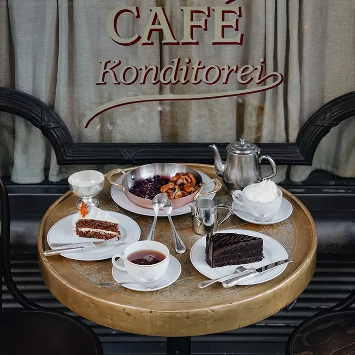 A round café table with two slices of cake, a bowl of dessert, whipped cream, a teapot, a teacup with tea, and silverware. “Café Konditorei” is written on the window behind the table.