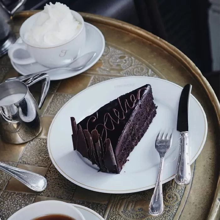 A slice of chocolate cake with decorative chocolate pieces and writing sits on a white plate with a fork and knife beside a cup of whipped cream and a small metal jug on a tray.