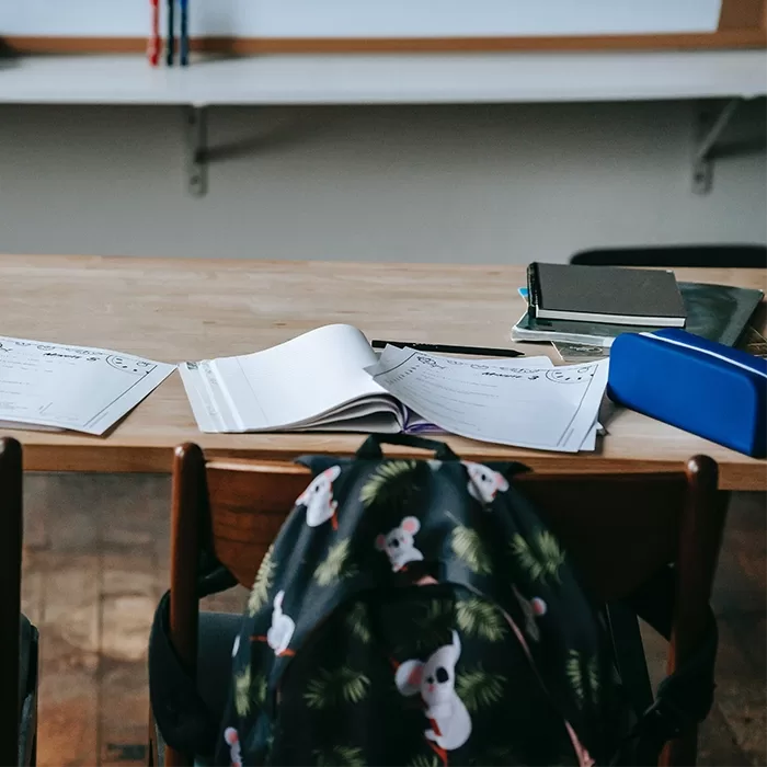 A wooden classroom desk with open notebooks, papers, a stack of books, a pencil case, and a backpack with a koala pattern hanging on the chair. The background shows a whiteboard and empty shelves.