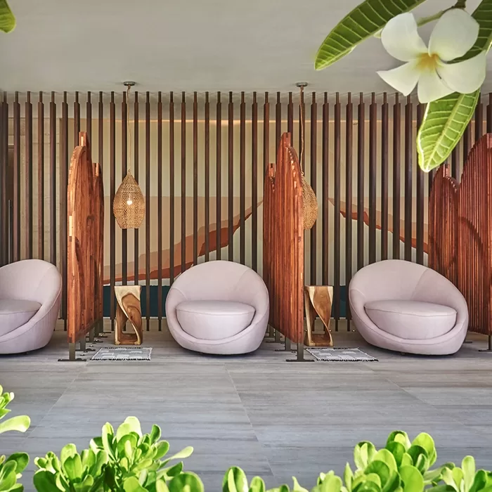 Modern lounge area with three round, light beige chairs separated by wooden dividers, woven pendant lights above, and green plants and white flowers framing the scene in the foreground.
