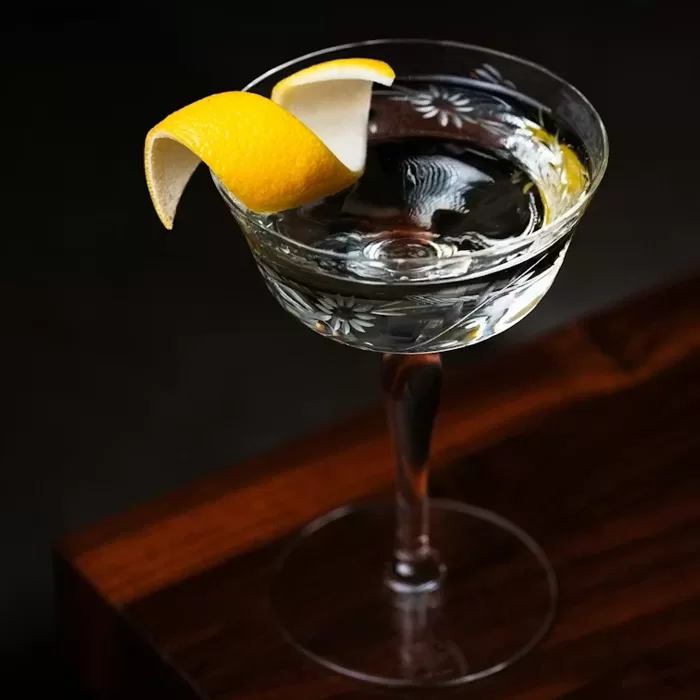 A clear cocktail in an etched glass sits on a dark wooden surface, garnished with a twisted lemon peel on the rim.