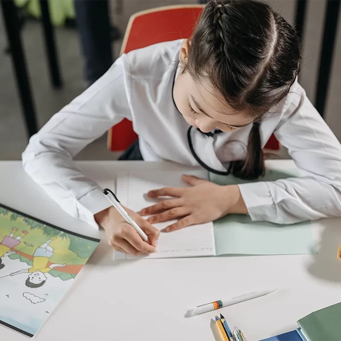 A young girl with braided hair sits at a desk, writing in a notebook. A colorful drawing and pens are on the table. She appears focused on her work, wearing a white shirt and sitting on a red chair.
