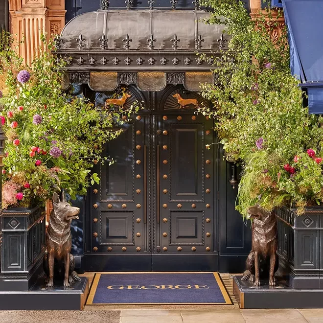 Ornate entrance with a black door adorned with golden studs, flanked by lush greenery and flowers. Bronze dog statues sit on either side, and the doormat reads GEORGE. A glass canopy with a metal frame shelters the doorway.