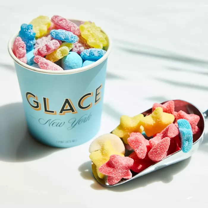 A blue cup labeled GLACE New York is filled with colorful gummy candies. Next to it, a metal scoop holds more gummies in various shapes and bright colors, all coated in sugar.
