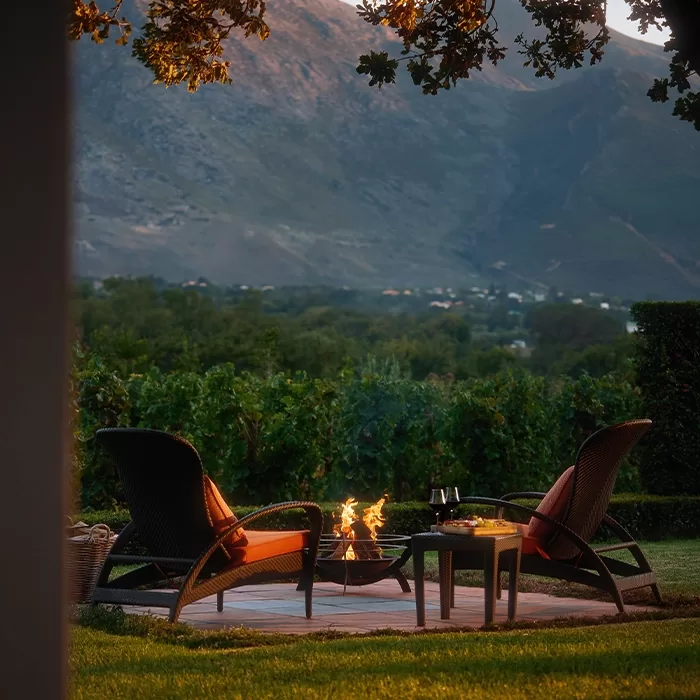 Two lounge chairs with red cushions face a small fire pit on a patio, surrounded by greenery and vineyards, with mountains in the background; a table holds two glasses of red wine and snacks.