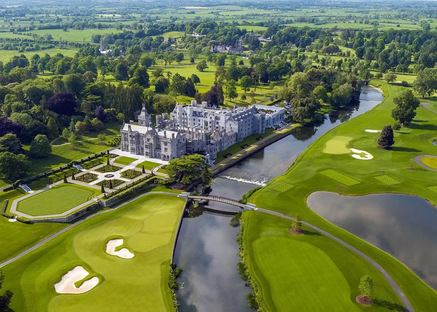 Aerial view of a large, historic castle-like mansion surrounded by lush green gardens, a moat, and expansive golf course with sand bunkers. The landscape is dotted with trees and stretches into the distance under a clear sky.