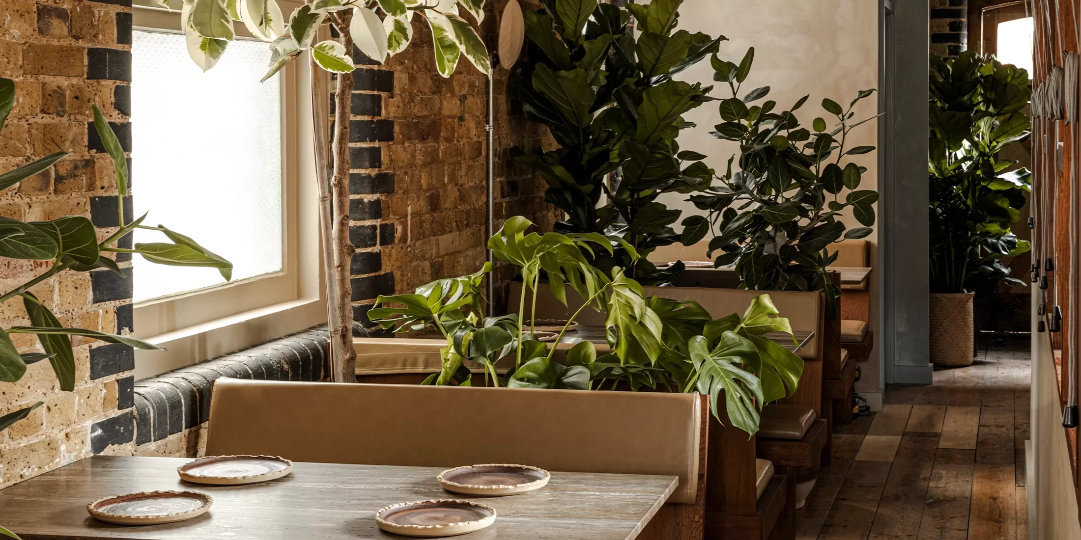 A cosy restaurant interior with wooden tables set with plates, tan cushioned booths, exposed brick walls, large windows, and lush green potted plants creating a warm, inviting atmosphere.