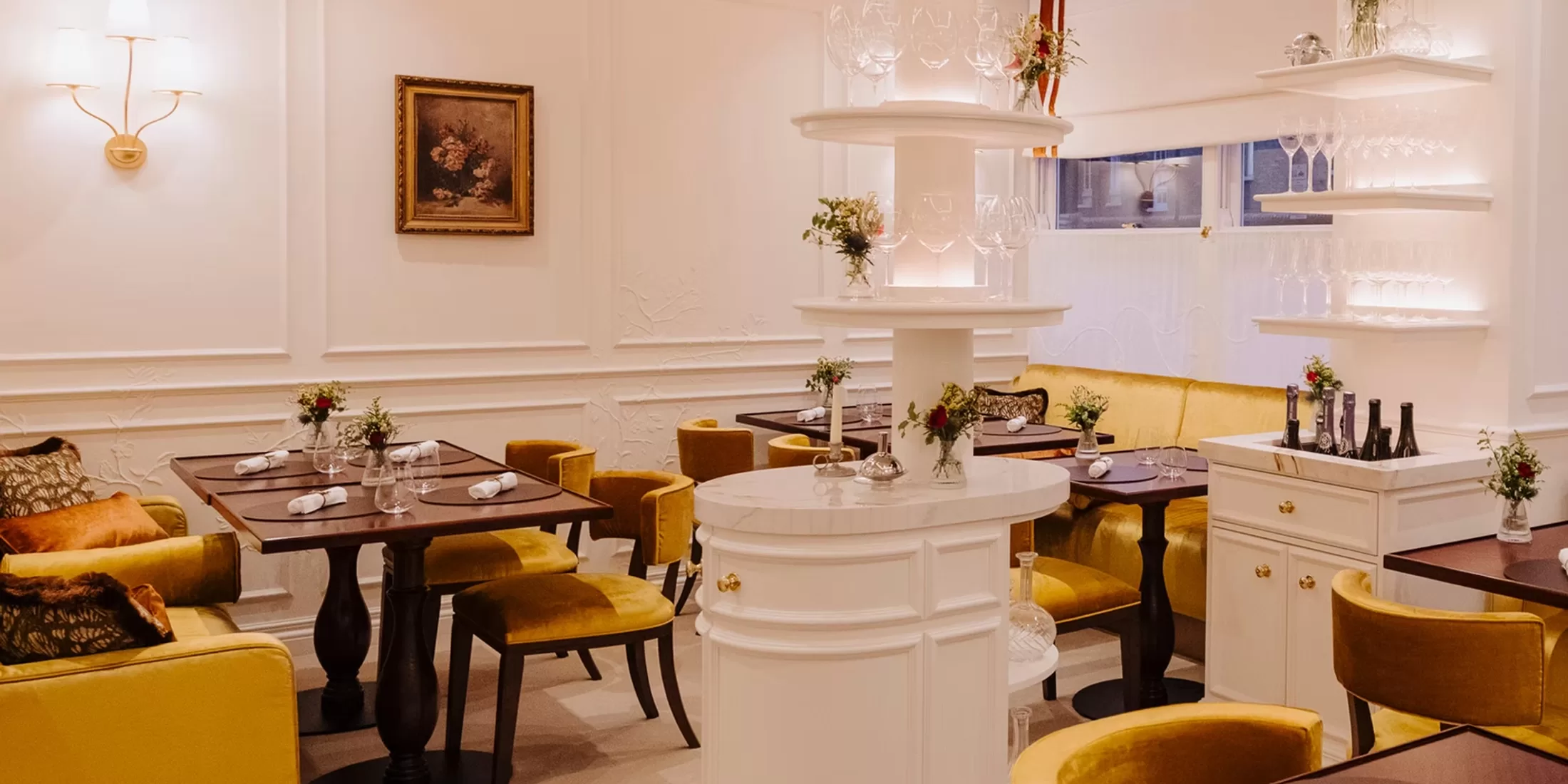Cozy restaurant interior with wooden tables, yellow velvet chairs, and benches. White walls, elegant decor, flowers on tables, a central white shelving unit with glassware, and a framed painting create a warm, inviting atmosphere.