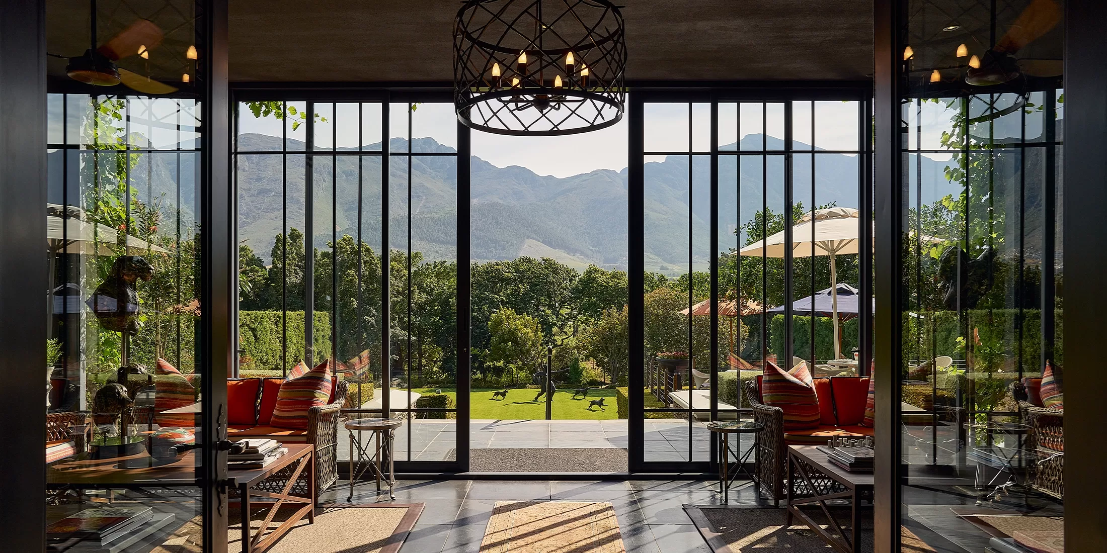 A luxurious lounge with glass doors opens to a lush garden, outdoor seating, and mountain views in the distance. The space features modern decor, striped pillows, and natural light filling the room.