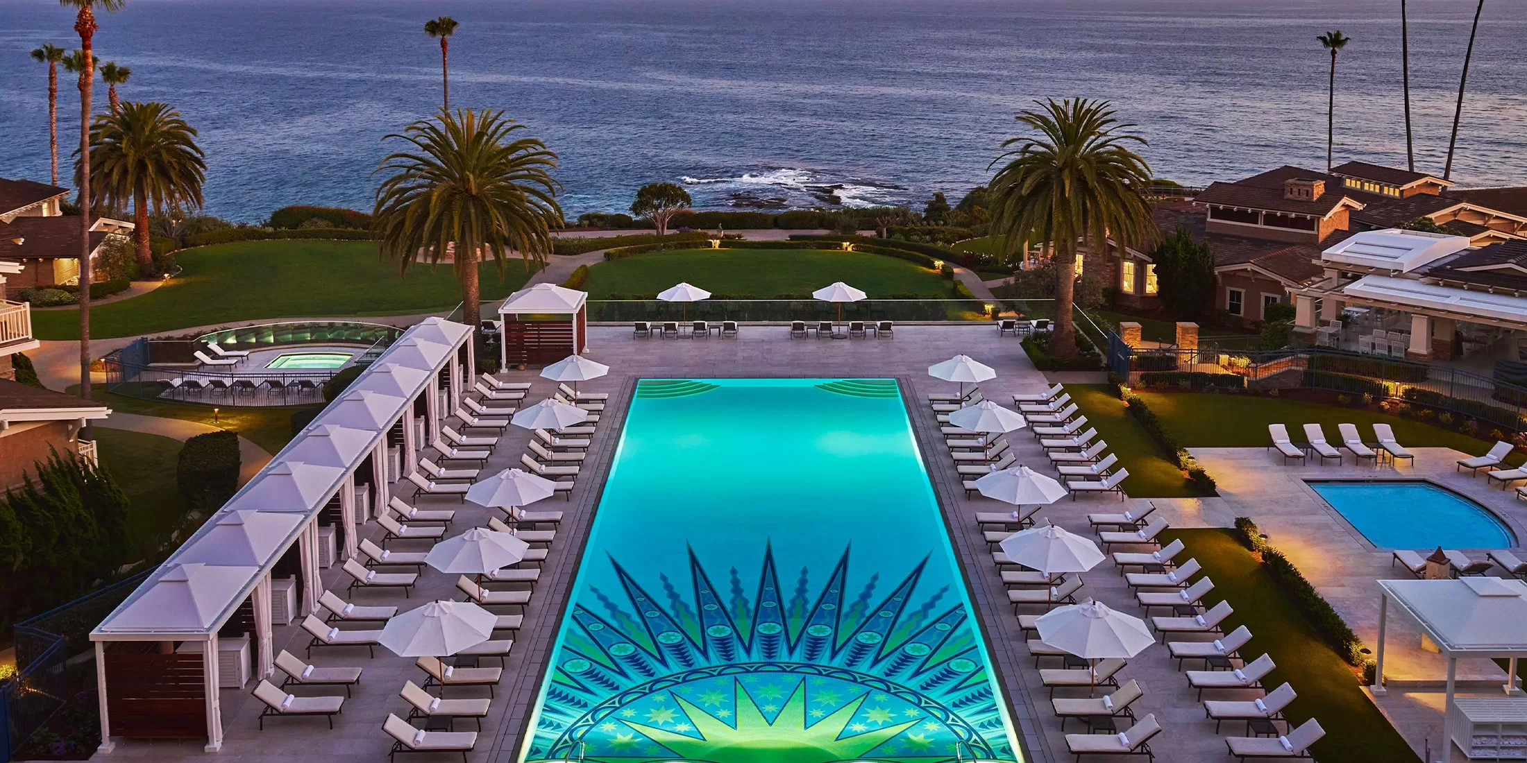 A luxurious outdoor pool with a sunburst design is surrounded by lounge chairs, umbrellas, palm trees, and cabanas, overlooking the ocean at sunset.
