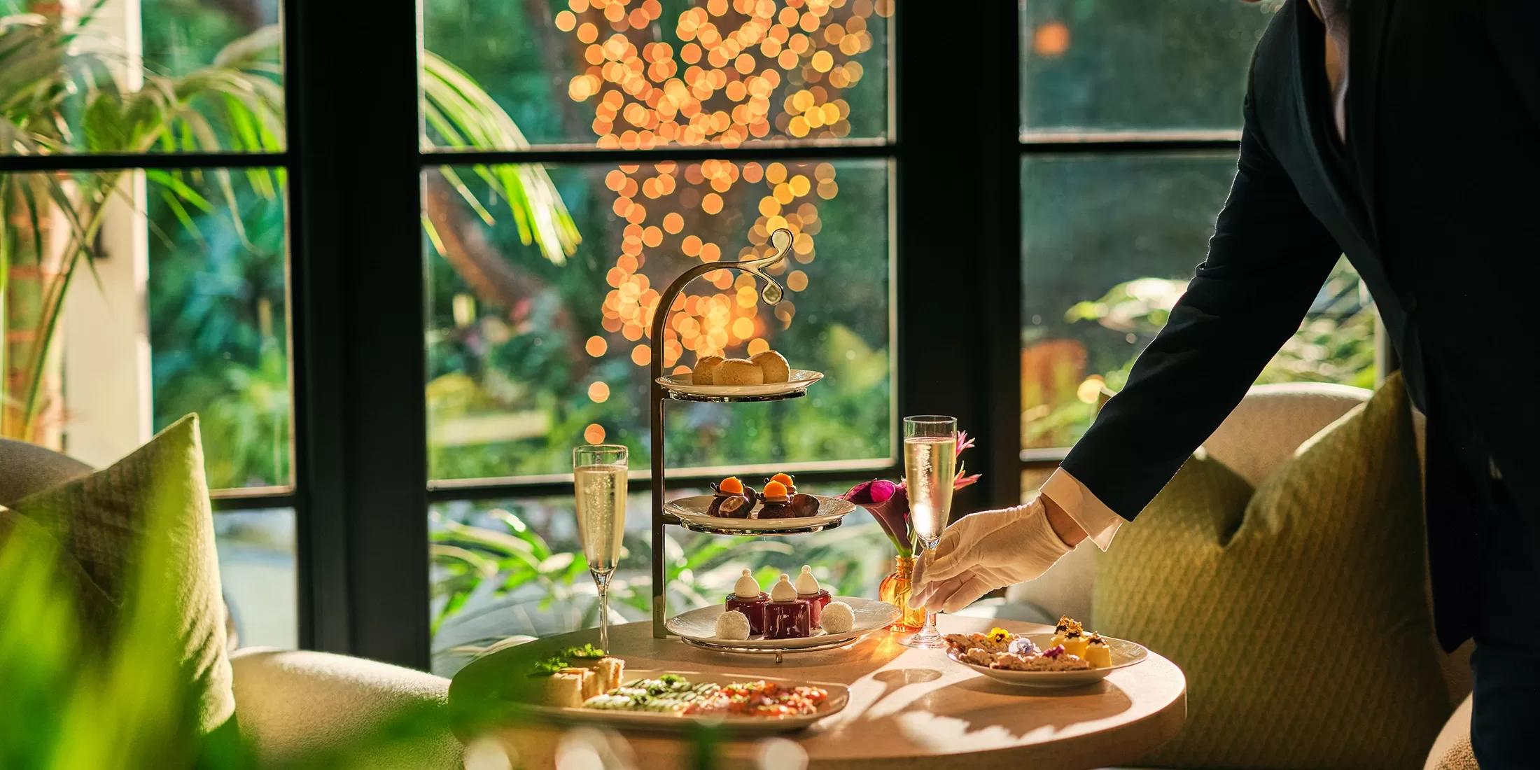 A person in formal attire serves afternoon tea, including pastries and sweets on a tiered tray, with a glass of champagne on a sunlit table by large windows overlooking a green garden with warm lights outside.