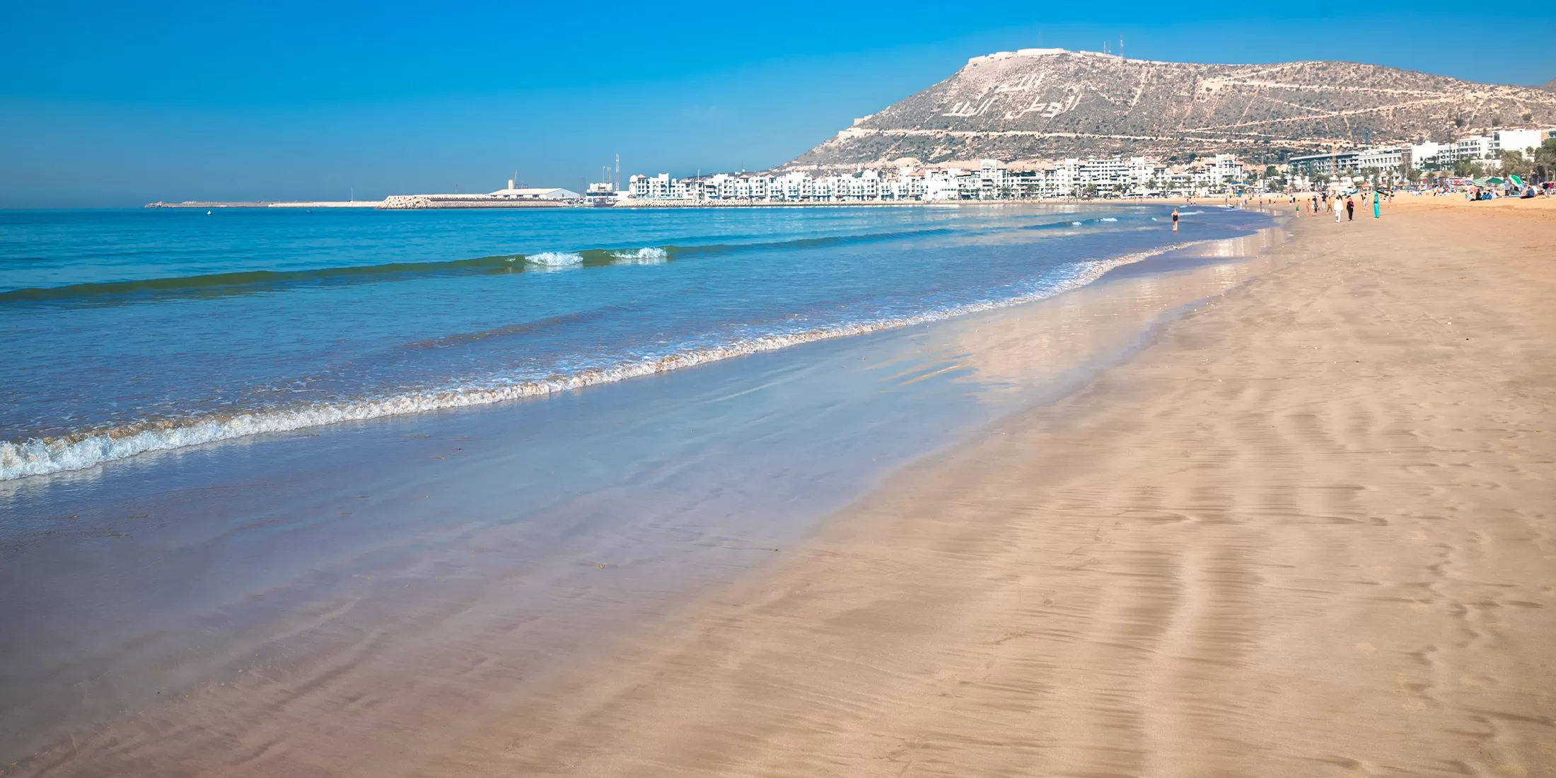 Sandy beach with gentle waves and a clear blue sky. In the distance, theres a mountain topped with a white structure and a cityscape with white buildings lining the shore. People are walking along the beach in Temuda Bay.