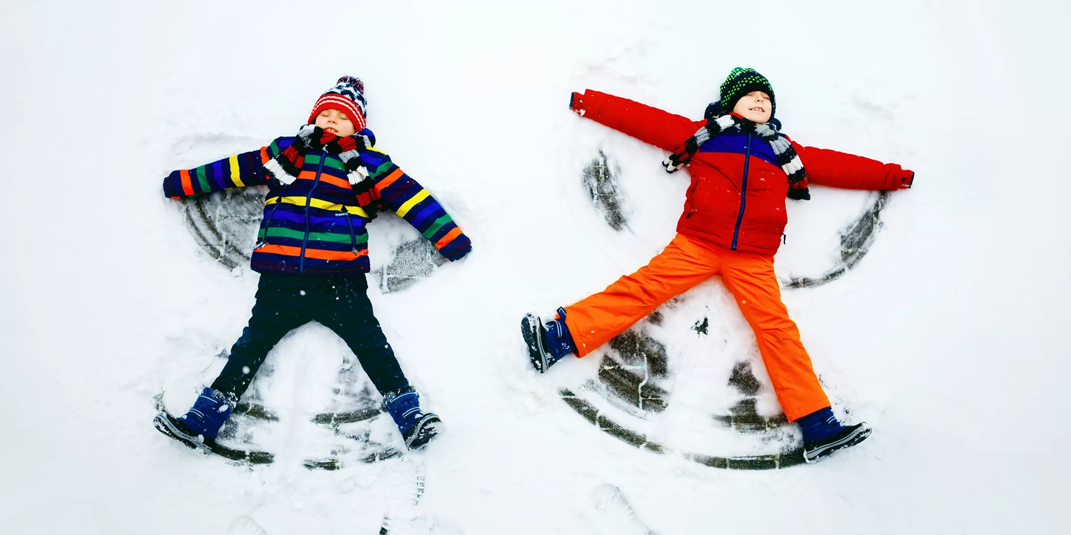 Two children dressed in colorful winter clothes lie on their backs making snow angels. The child on the left wears a striped jacket, and the one on the right wears an orange and red outfit. Both smile joyfully against the snowy backdrop.