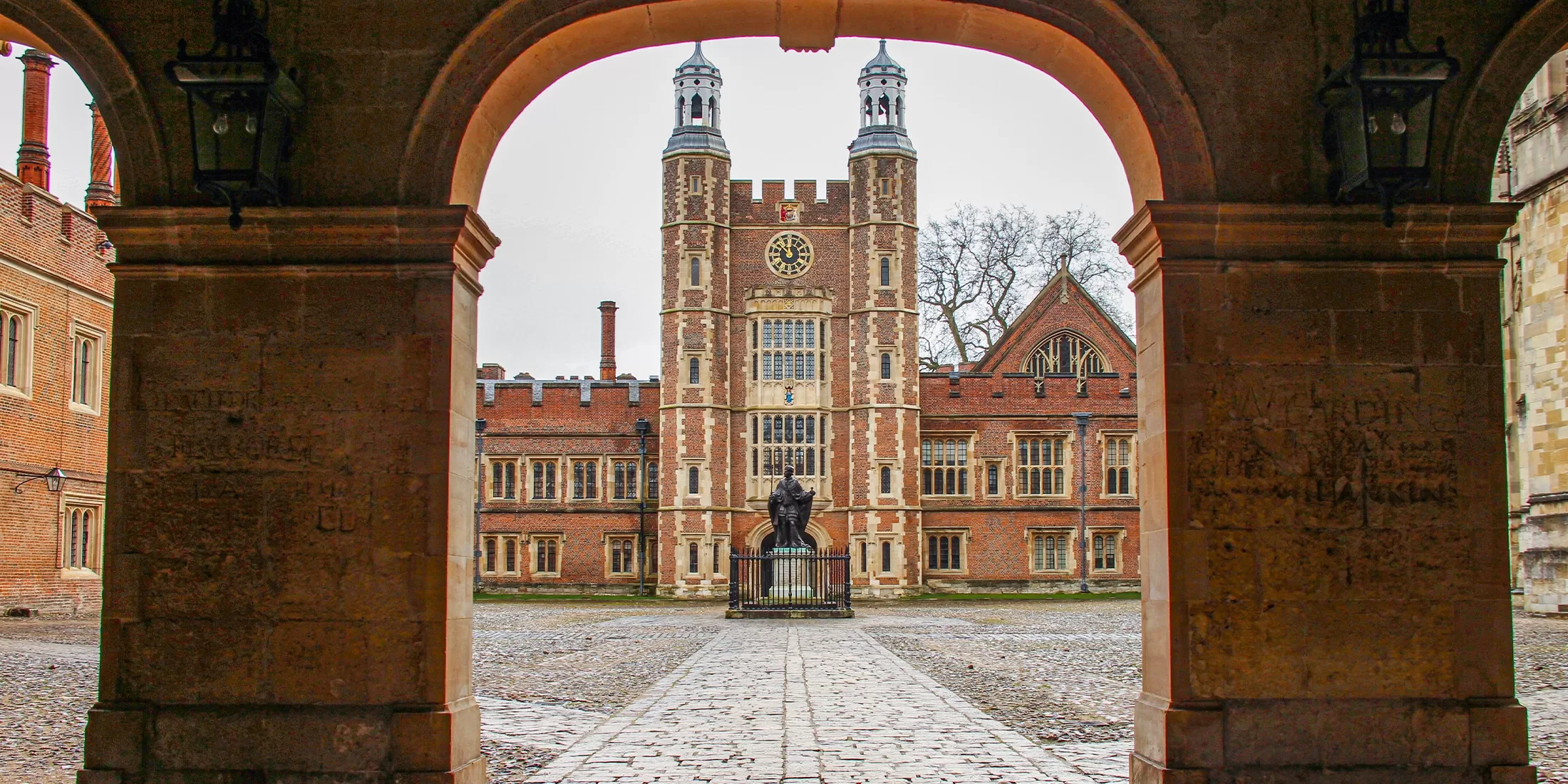 View through an archway of a historic brick building with a clock tower, central statue, cobblestone courtyard, and gothic architectural details on a cloudy day.