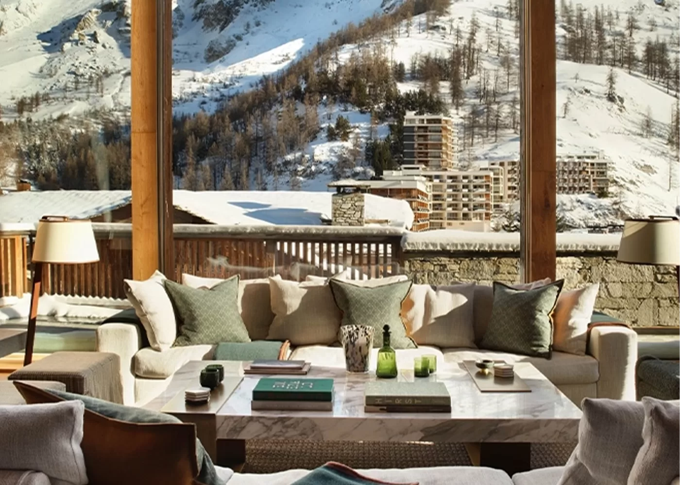 Cozy living room with a large wooden-framed window offering a scenic view of snow-covered mountains and trees. The room features comfortable beige sofas adorned with cushions around a central coffee table with decorative items.