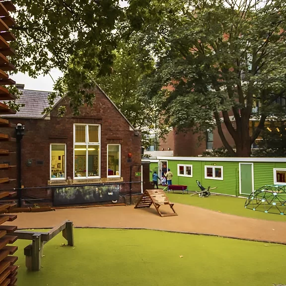 A playground with green artificial turf, climbing structures, a brown ramp, and a balance beam. Behind, there is a brick building with large windows and a green modular classroom. Trees provide shade overhead.