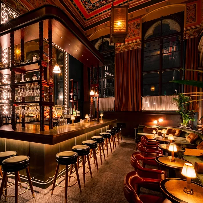 A stylish, warmly lit bar with tall stools lined up along the counter, cozy armchairs, round tables with small lamps, rich wood accents, and ornate ceiling details in an elegant, inviting setting.
