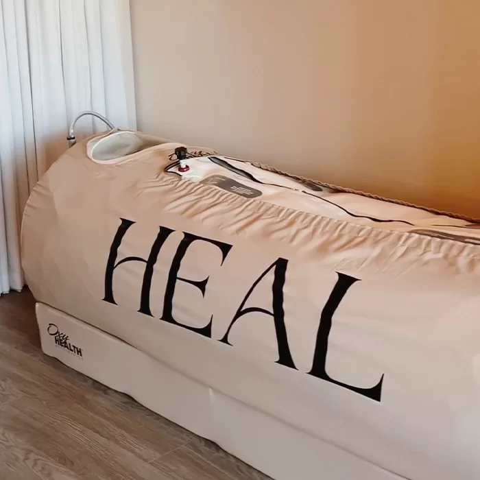 A beige hyperbaric oxygen therapy chamber labeled HEAL sits in a room with light wooden floors and cream-colored curtains.