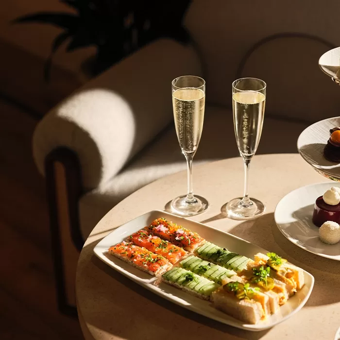 A table with two glasses of sparkling wine and a plate of assorted finger sandwiches, including fillings of cucumber, smoked salmon, and egg, bathed in warm sunlight.