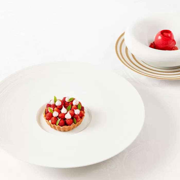 A small tart topped with strawberries, dollops of cream, and mint leaves sits on a large white plate. In the background, a bowl holds a bright red dessert, possibly sorbet, on a white tablecloth.