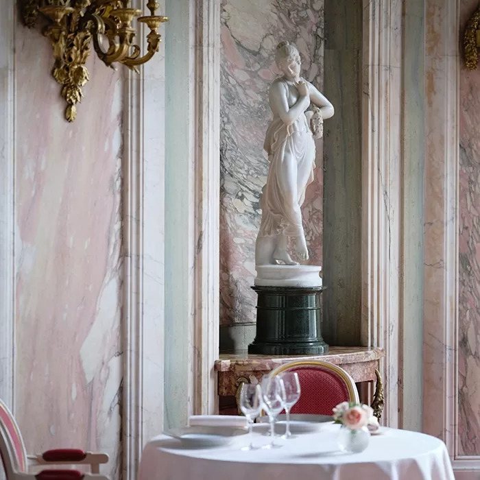 A marble statue of a woman stands in an ornate alcove with pink marble walls. In front, a round table is set with wine glasses, plates, red chairs, and a small bouquet of roses. Gold wall sconces are visible on the left.