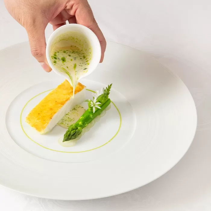 A hand pours a creamy green herb sauce over a rectangular piece of fish and an asparagus spear, artfully plated on a white dish with a ring of green sauce.