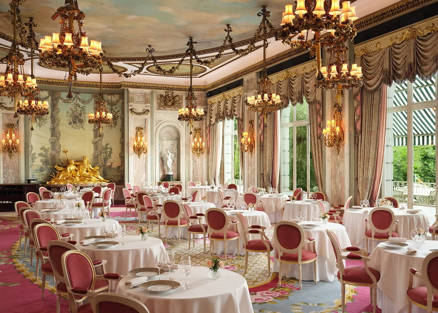 A grand, elegant dining room with ornate chandeliers, large windows, pink and gold chairs, round tables with white cloths, lavish drapes, and a decorative gold sculpture as a centerpiece on one side.