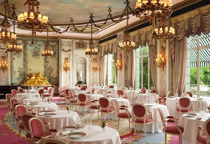 Elegant, ornate dining room with pink chairs, round tables set with white linens, chandeliers, large windows, and detailed gold decor. Classical paintings and statues adorn the walls, creating a luxurious atmosphere.