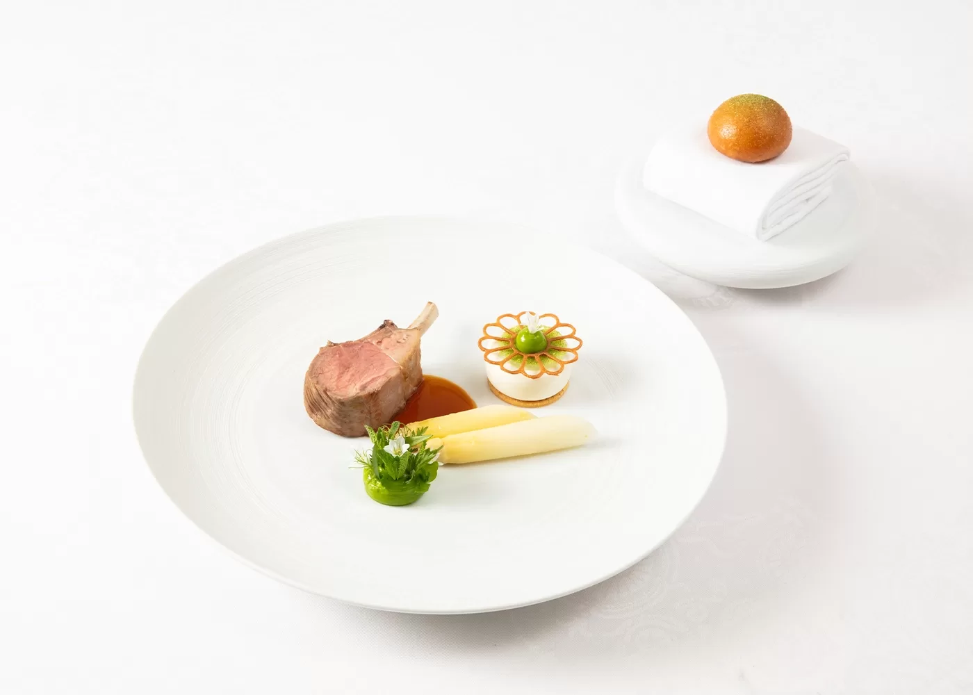 A gourmet dish featuring a rack of lamb with sauce, two white asparagus spears, broccoli, and a decorative vegetable arrangement, served on a white plate. In the background, a mandarin orange sits atop a folded white napkin.