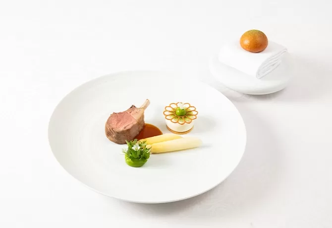 A gourmet dish featuring a lamb chop on sauce, an artfully arranged potato flower, green garnish, and two asparagus spears on a white plate. A round orange pastry and napkin are on a separate plate in the background.