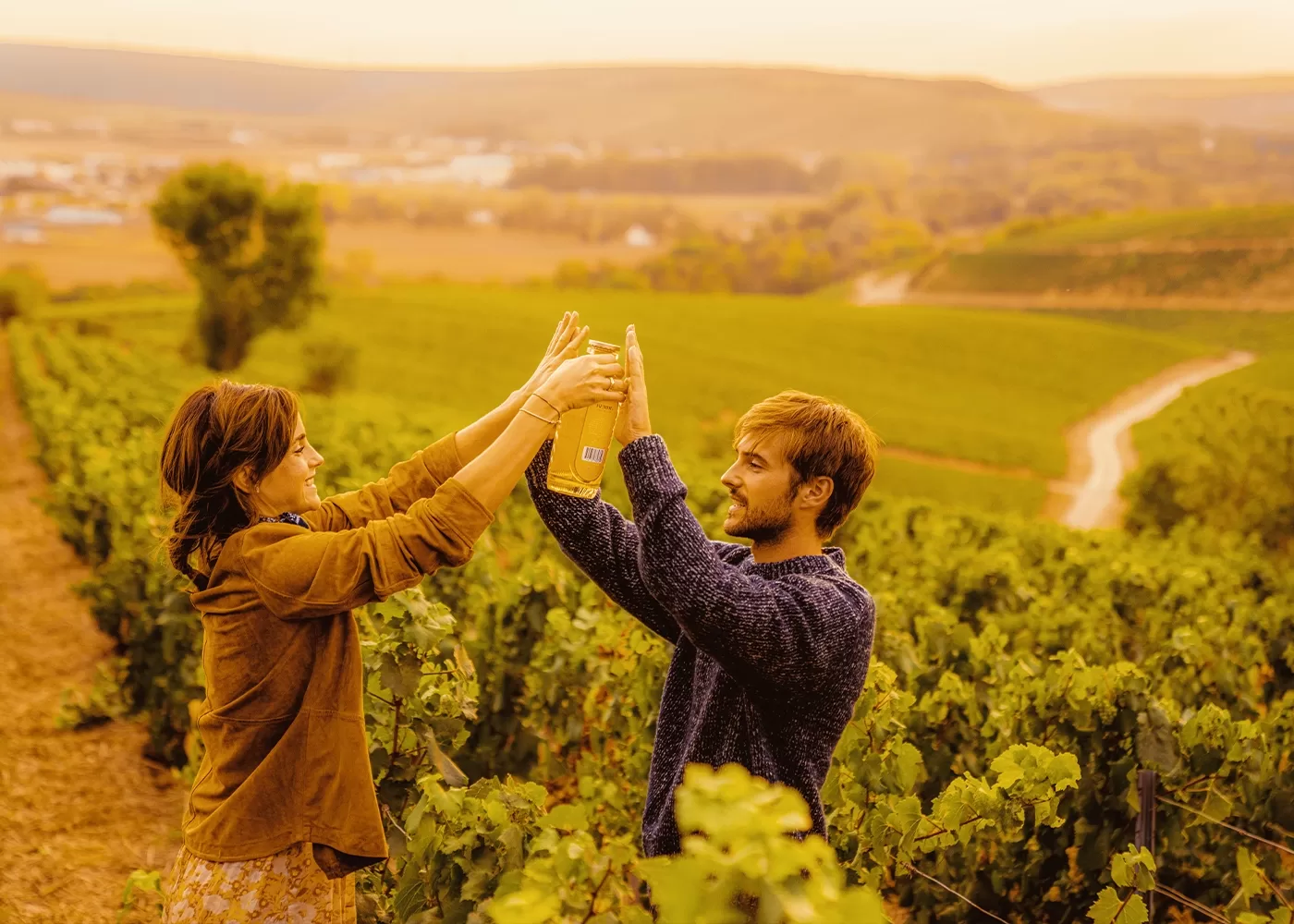 Two people stand among lush green vineyard rows, both smiling and reaching up to hold a wine bottle together, with golden sunlight and rolling hills in the background.