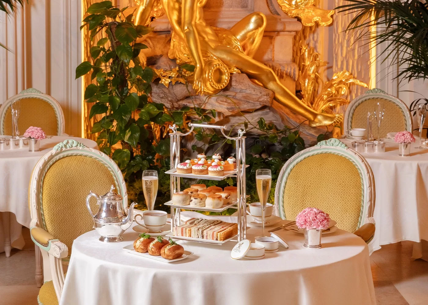 An elegant afternoon tea setup with tiered trays of pastries and sandwiches, a silver teapot, champagne glasses, and pink flowers on a round table, set in a luxurious room with gold decor and classic chairs.