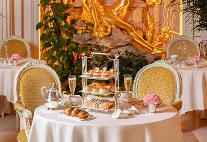 A lavish afternoon tea setup with elegant tables, tiered trays of pastries and sandwiches, teacups, champagne glasses, and pink flowers, set in a luxurious room with gold statues and ornate furnishings.