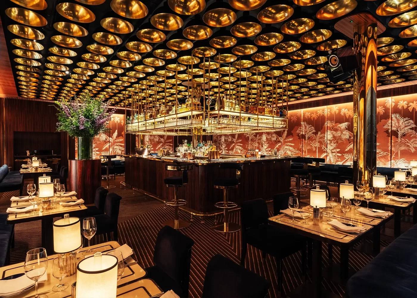 Elegant restaurant interior with round golden ceiling lights, a central bar surrounded by stools, tables set with lamps and glassware, dark tones, floral wall patterns, and a large vase of purple flowers.
