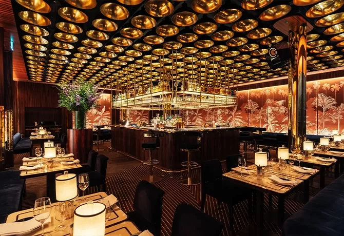 Elegant restaurant interior with gold ceiling accents, soft table lamps, dark wood furniture, and a central bar surrounded by neatly set dining tables. Warm lighting and decorative wall panels create a luxurious, inviting atmosphere.