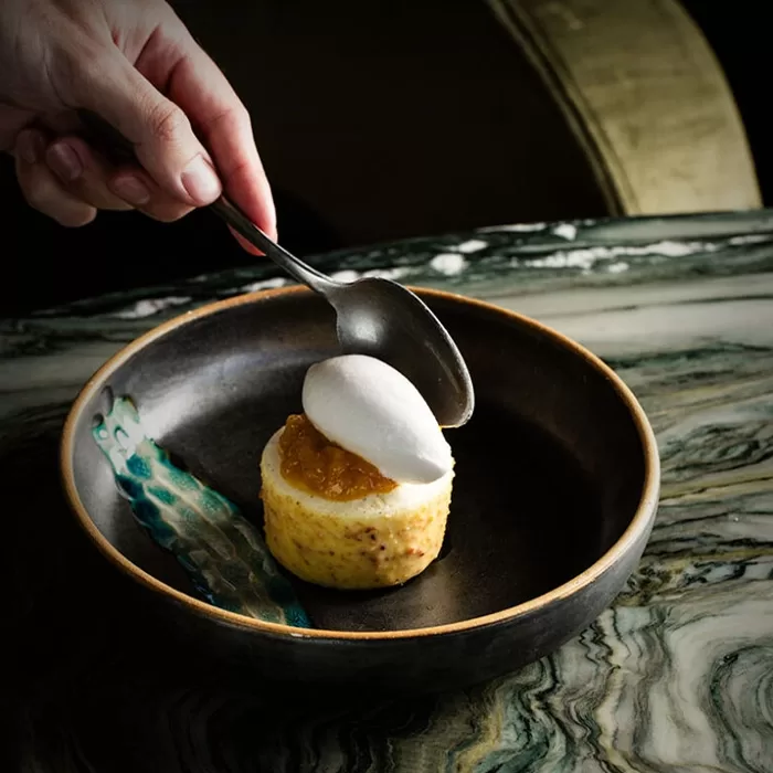 A hand holds a spoon above a dessert topped with white cream and a yellow fruit compote, served on a dark plate with a decorative green streak, on a marbled tabletop.