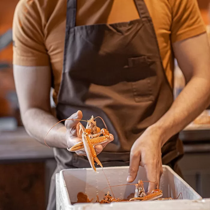 A person wearing a brown apron and orange shirt holds a raw lobster above a white container filled with seafood, possibly in a kitchen or restaurant setting.