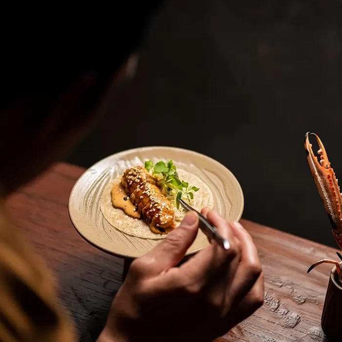 A person holds a fork, preparing to eat a gourmet dish featuring a glazed, garnished piece of seafood on a beige plate, with greens and sauce artfully arranged on a wooden table.