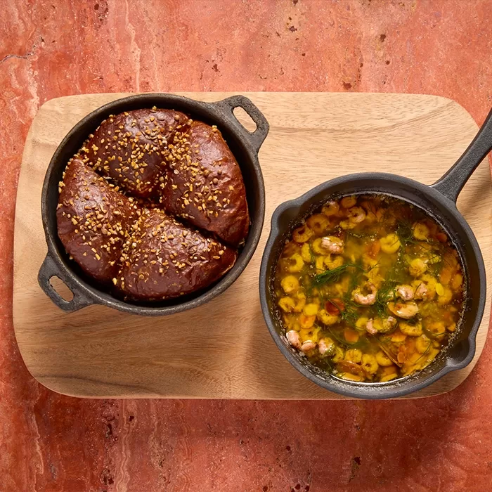 Two small cast iron pans on a wooden board: one contains three pieces of bread topped with sesame seeds, and the other holds shrimp cooked in garlic and oil, garnished with herbs.